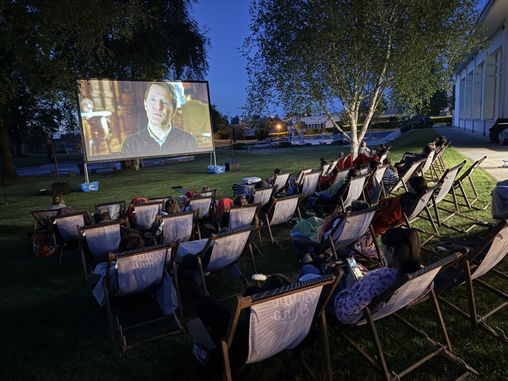 Menschen sitzen auf Liegestühlen auf einer Wiese und sehen sich in der Abenddämmerung einen Film an, der auf eine große Leinwand projiziert wird. Der Bereich ist von Bäumen umgeben, und rechts ist ein Gebäude mit großen Fenstern zu sehen.