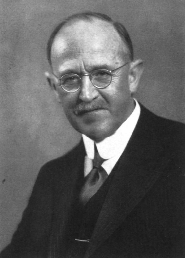 Ein älterer Mann mit Schnurrbart und runder Brille, der einen dunklen Anzug, ein weißes Hemd und eine Krawatte trägt, posiert für ein formelles Schwarz-Weiß-Porträt vor einem schlichten Hintergrund.