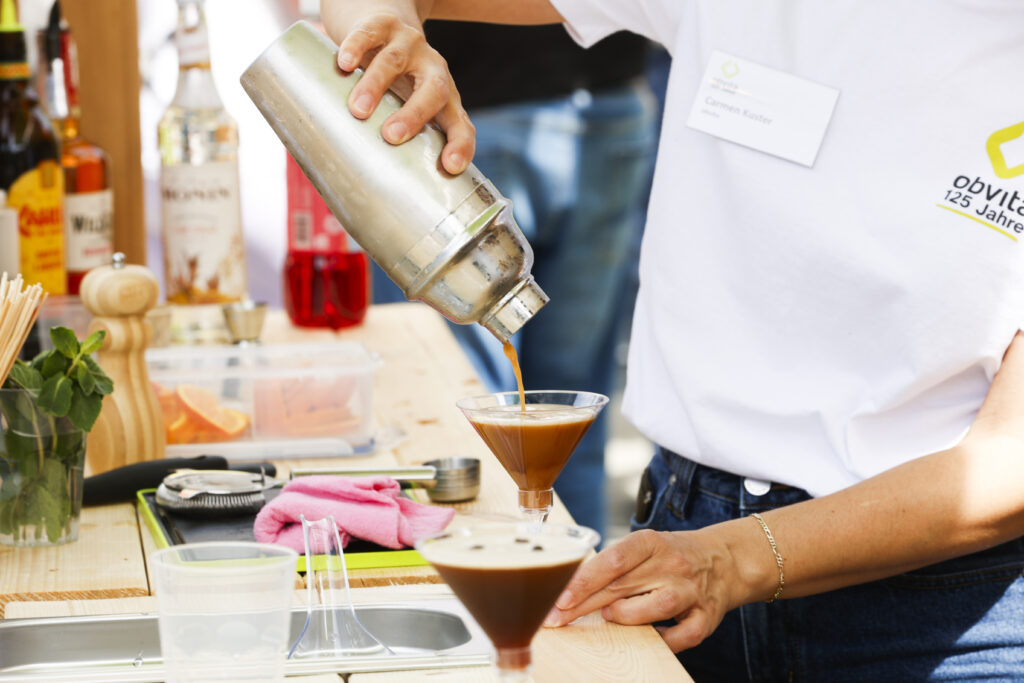 Eine Person in einem weißen Hemd gießt einen Cocktail aus einem Shaker in ein Martiniglas auf einem Holztisch, umgeben von Flaschen, einer Pflanze und Barutensilien in hellem Tageslicht.