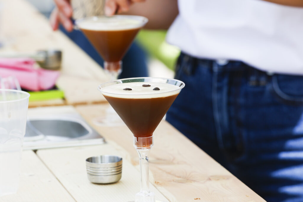 Nahaufnahme eines Espresso Martini, garniert mit Kaffeebohnen in einem Martiniglas, mit einer daneben stehenden Person und einem weiteren Getränk, das im Hintergrund auf einem Holztisch zubereitet wird.