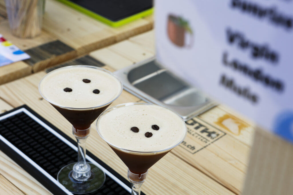 Auf einem hölzernen Bartresen stehen zwei Espresso-Martinis in Cocktailgläsern, die jeweils mit drei Kaffeebohnen bestückt sind. Im Hintergrund sind die Barausstattung und ein teilweise sichtbares Schild mit der Speisekarte zu sehen.