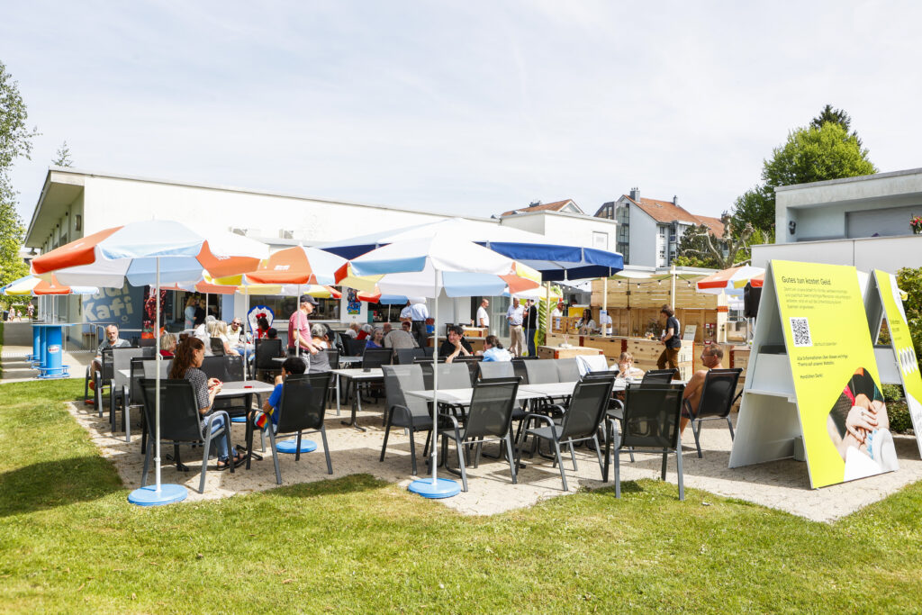 Menschen sitzen an Tischen im Freien unter bunten Sonnenschirmen in einem sonnigen Café. Im Hintergrund sind Gebäude zu sehen und im Vordergrund ein großes gelbes Schild. Die Atmosphäre ist lebhaft und entspannt.