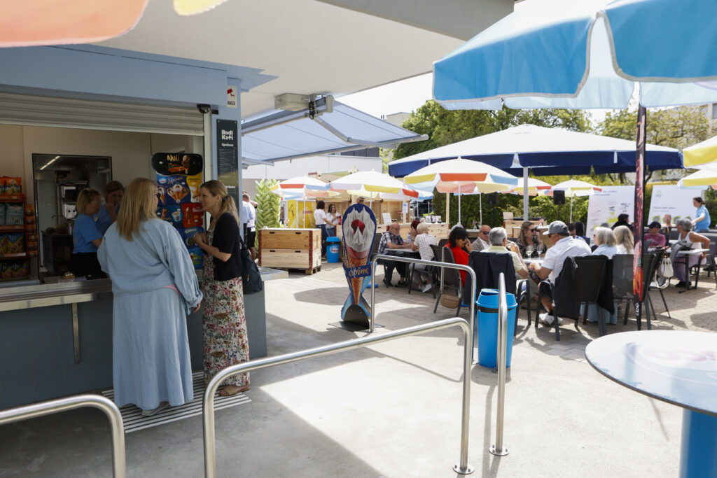 Die Leute bestellen Essen an einer Snackbar im Freien, während andere an Tischen unter bunten Schirmen sitzen und an einem sonnigen Tag essen und sich unterhalten. Die Atmosphäre ist lebhaft und entspannt.
