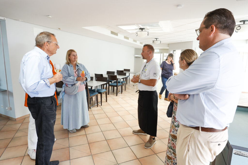 Eine Gruppe von fünf Erwachsenen steht in einem Kreis in einem hellen Raum mit Fliesenboden, unterhält sich und lächelt. Einige sind leger gekleidet, andere tragen Business-Kleidung. Leere Stühle stehen an der Wand hinter ihnen, und eine weitere Person steht im Hintergrund.