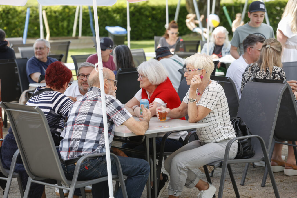 Eine Gruppe von Menschen unterschiedlichen Alters sitzt an einem sonnigen Tag im Freien an Tischen, unterhält sich und genießt Getränke bei einem geselligen Beisammensein. Einige Menschen stehen im Hintergrund, während hinter ihnen üppiges Grün zu sehen ist.