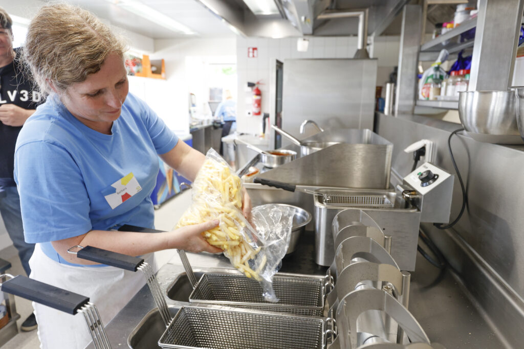 Eine Frau in einem blauen Hemd schüttet eine Tüte mit rohen Pommes frites in einen Frittierkorb in einer Großküche, um sie zu frittieren.