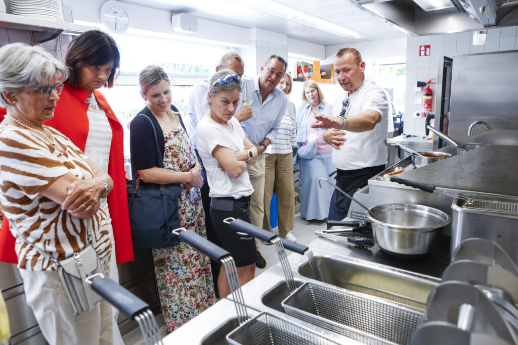 Ein Chefkoch erklärt einer Gruppe aufmerksamer Erwachsener in einer modernen, gut beleuchteten Küche die Küchengeräte. Im Vordergrund sind mehrere Friteusen und Töpfe zu sehen. Die Gruppe steht dicht beieinander, hört zu und beobachtet.