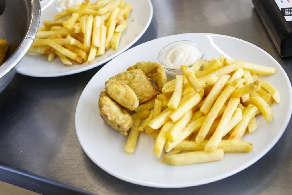 Ein weißer Teller mit goldenen Pommes frites, drei Stücken paniertem Fisch und einer kleinen Portion cremiger Dip-Sauce auf einer Edelstahlfläche. Ein weiterer ähnlicher Teller ist teilweise im Hintergrund zu sehen.