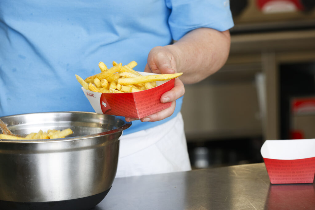 Eine Person in einem blauen Hemd hält eine rote Pappschachtel mit Pommes frites in der Hand und schöpft sie aus einer Metallschüssel auf einer Edelstahltheke in einer Restaurantküche.
