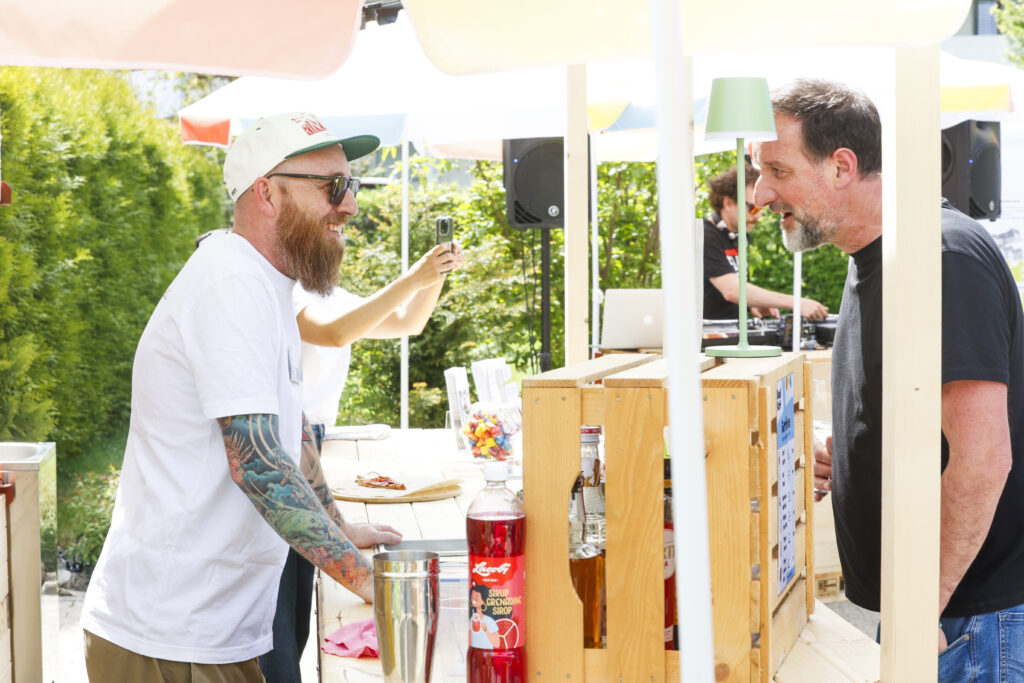 Ein bärtiger Mann mit Tattoos, der eine Sonnenbrille und eine weiße Kappe trägt, lächelt, während er sich mit einem anderen Mann an einer Bar im Freien unterhält. Auf dem Tresen stehen Flaschen und ein Shaker, und im Hintergrund macht eine Frau ein Foto.