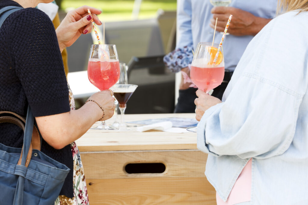 Zwei Personen mit Gläsern mit rosa Cocktails und gestreiften Strohhalmen stehen an einem Holztisch im Freien. Eine weitere Person im Hintergrund hält ein dunkles Getränk in einem Cocktailglas. Alle sind leger gekleidet.