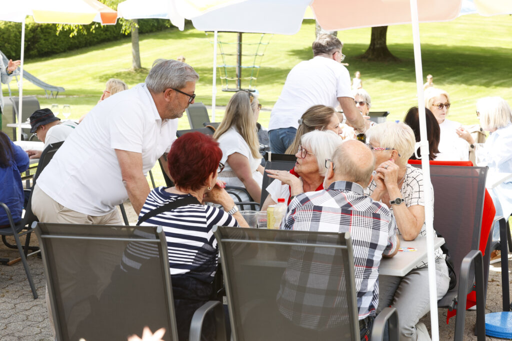Eine Gruppe älterer Menschen sitzt an Tischen im Freien unter Sonnenschirmen, unterhält sich und genießt den sonnigen Tag in einer parkähnlichen Umgebung. Ein Mann steht und lehnt sich in Richtung einer sitzenden Gruppe, während sich andere in der Nähe unterhalten und entspannen.
