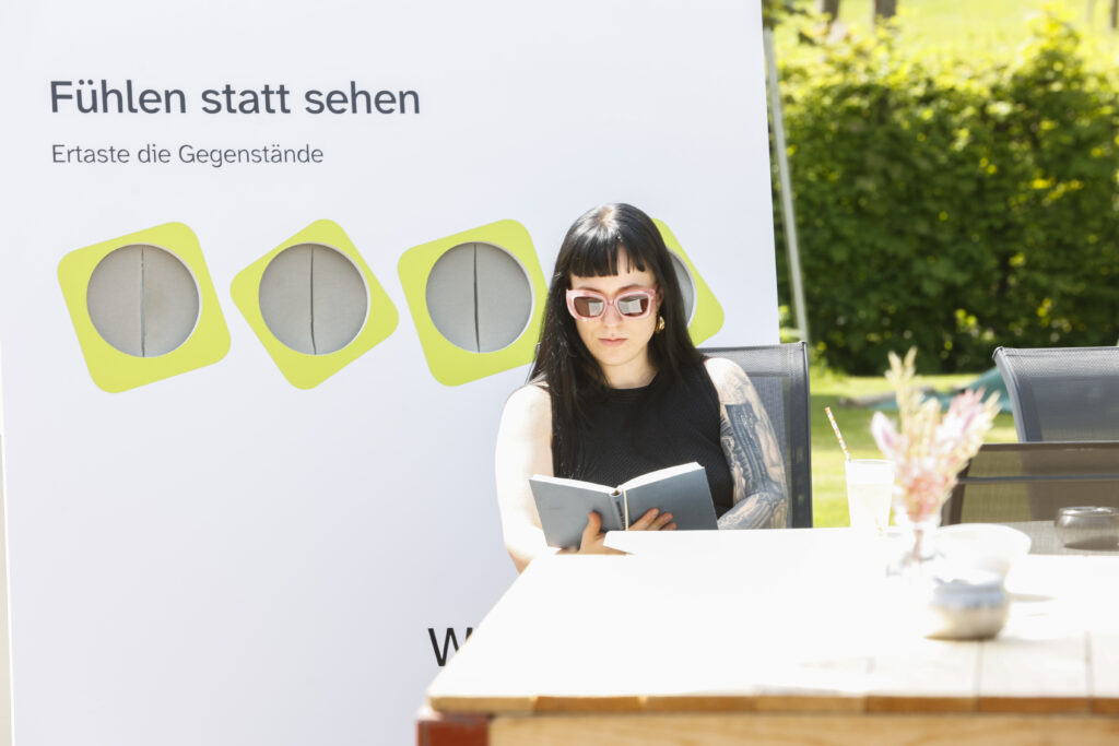 Eine Frau mit dunklem Haar und Sonnenbrille sitzt im Freien an einem Tisch und liest ein Buch. Hinter ihr steht ein weißes Schild in deutscher Sprache mit den Worten "Fühlen statt sehen" und "Errate die Gegenstände", mit drei tastbaren Kreisen.