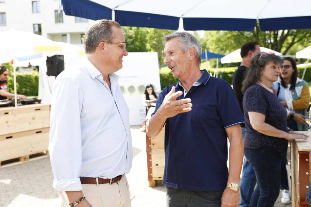 Zwei Männer stehen draußen unter einem großen weiß-blauen Vordach und unterhalten sich bei einer gesellschaftlichen Veranstaltung. Im Hintergrund sind Menschen und Holzbuden an einem sonnigen Tag zu sehen.