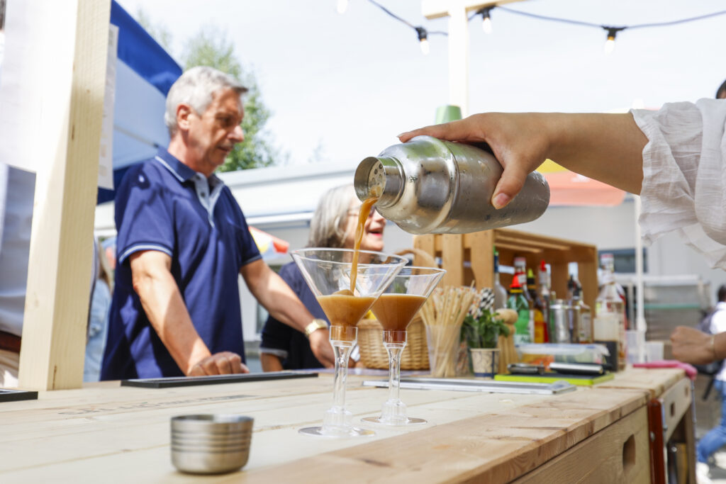 Ein Barkeeper gießt Cocktails aus einem Shaker in zwei Martini-Gläser an einer Bar im Freien, während zwei Personen auf der anderen Seite des Tresens stehen. Die Szene ist hell, lässig und lebendig.
