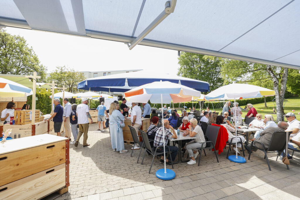 Menschen sind im Freien unter bunten Sonnenschirmen versammelt, sitzen an Tischen und gehen umher. Die Szene scheint ein lebhaftes gesellschaftliches Ereignis am Tag zu sein, möglicherweise ein Jahrmarkt oder eine Versammlung, mit Bäumen und grünem Gras im Hintergrund.