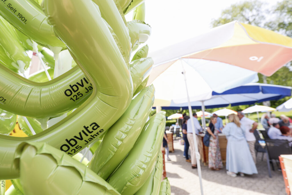 Grüne aufblasbare Objekte mit der Aufschrift "obvita 125 Jahre" stehen im Vordergrund. Im Hintergrund stehen Menschen und unterhalten sich unter bunten Regenschirmen bei einer Veranstaltung im Freien.