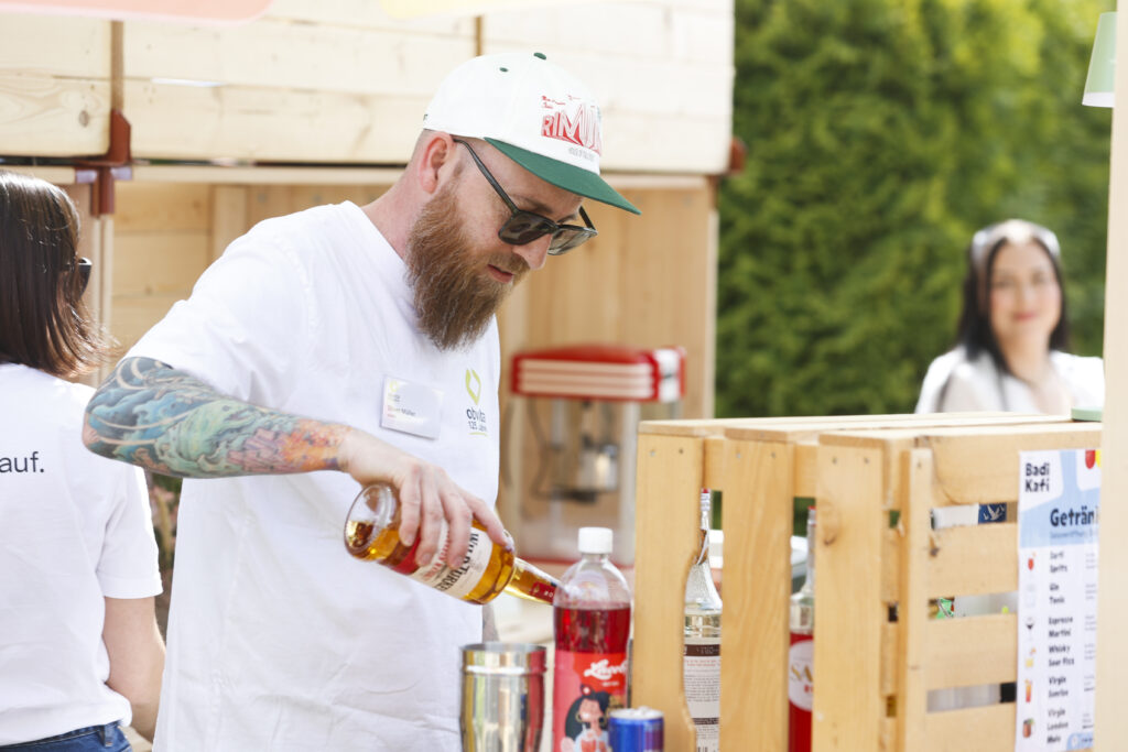 Ein bärtiger Mann mit Sonnenbrille und Mütze gießt eine Flasche Schnaps in einen Metallshaker an einem Stand im Freien, umgeben von verschiedenen Getränkeflaschen, mit Menschen und Grünflächen im Hintergrund.