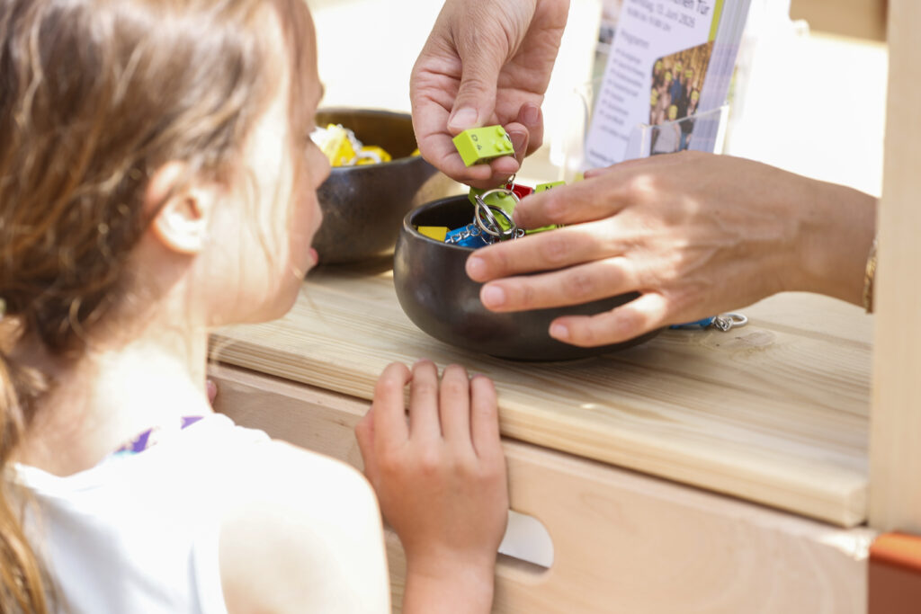 Ein junges Mädchen steht an einer Theke, während eine erwachsene Hand ein kleines grünes Spielzeug aus einer mit buntem Schmuck gefüllten Schale auswählt. Eine weitere Schale und ein Flyer sind ebenfalls auf der Holzoberfläche zu sehen.