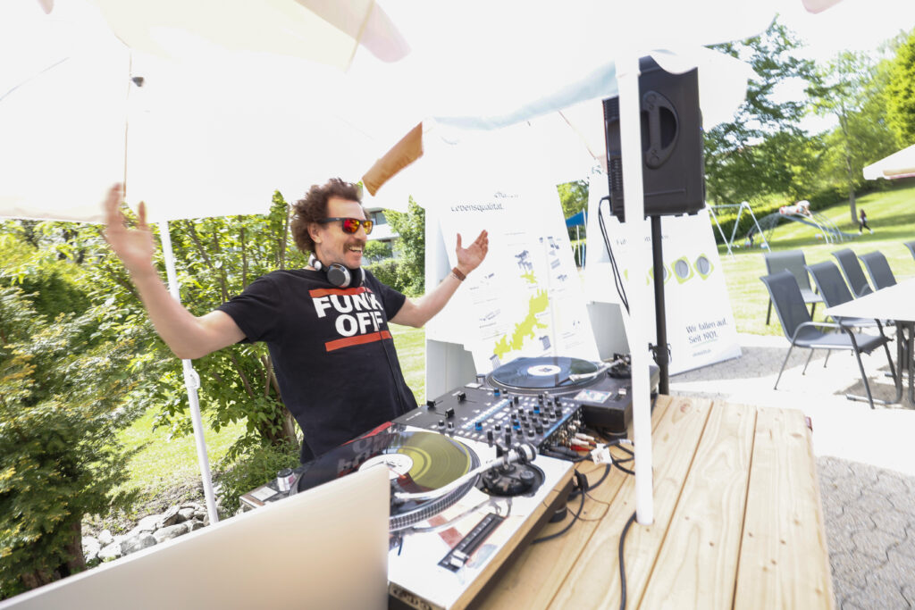 Ein DJ mit Sonnenbrille, Kopfhörern und einem "FUNK OFF"-T-Shirt spielt im Freien Musik auf Plattenspielern und lächelt mit erhobenen Armen. Im Hintergrund sind Terrassenstühle und Grünpflanzen zu sehen.