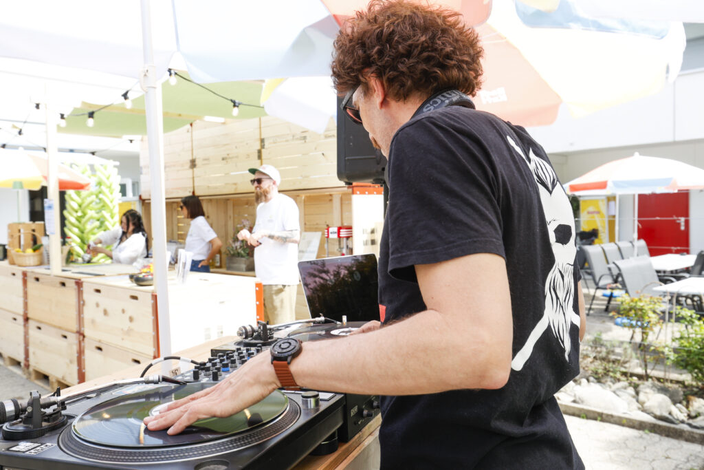 Ein DJ mit lockigem Haar mischt bei einer Veranstaltung im Freien Musik auf Plattentellern. In der Nähe treffen sich Leute, und bunte Sonnenschirme spenden Schatten über den Lounge-Sitzplätzen in einer sonnigen, entspannten Atmosphäre.