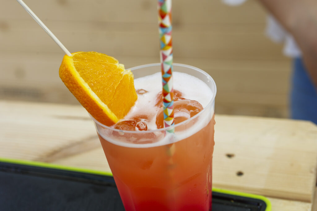 Ein kalter orange-rosa Cocktail mit Eis, garniert mit einer Orangenscheibe und einem bunt gemusterten Strohhalm, steht auf einer Holzfläche.