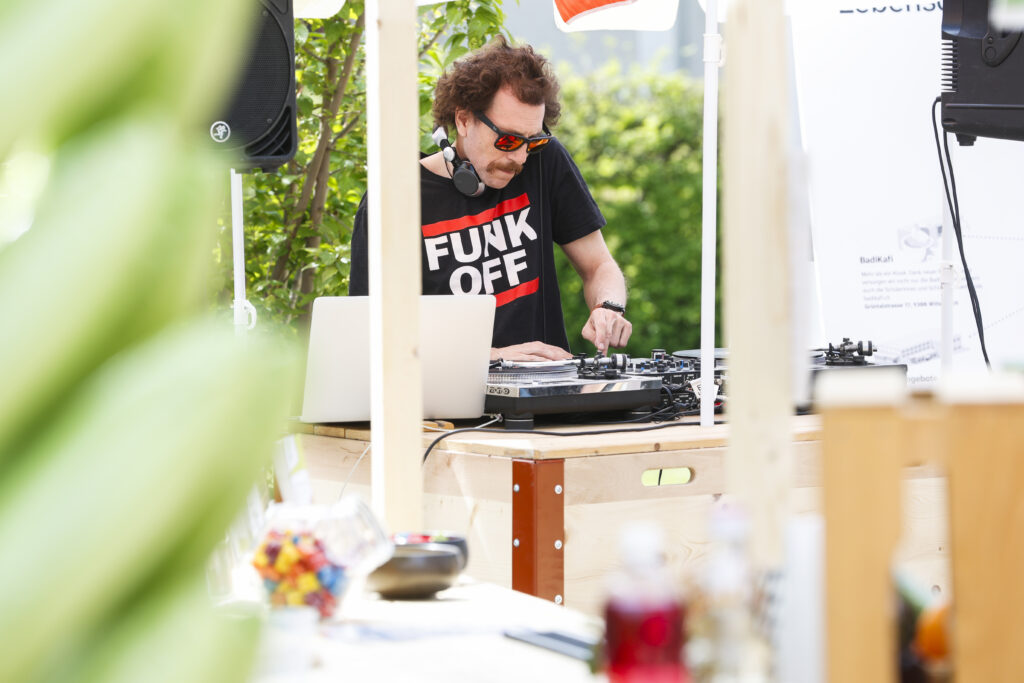 Ein DJ mit Sonnenbrille und "FUNK OFF"-T-Shirt mischt Musik auf einem Plattenteller im Freien, umgeben von Holzkonstruktionen und Grünpflanzen, mit einem Laptop und verschiedenen Gegenständen auf dem Tisch.