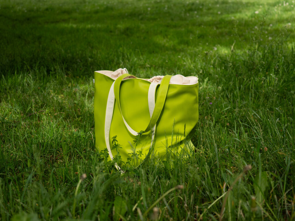 Eine leuchtend grüne Tragetasche mit beigen Henkeln sitzt auf saftig grünem Gras in einer sonnenbeschienenen Umgebung im Freien.