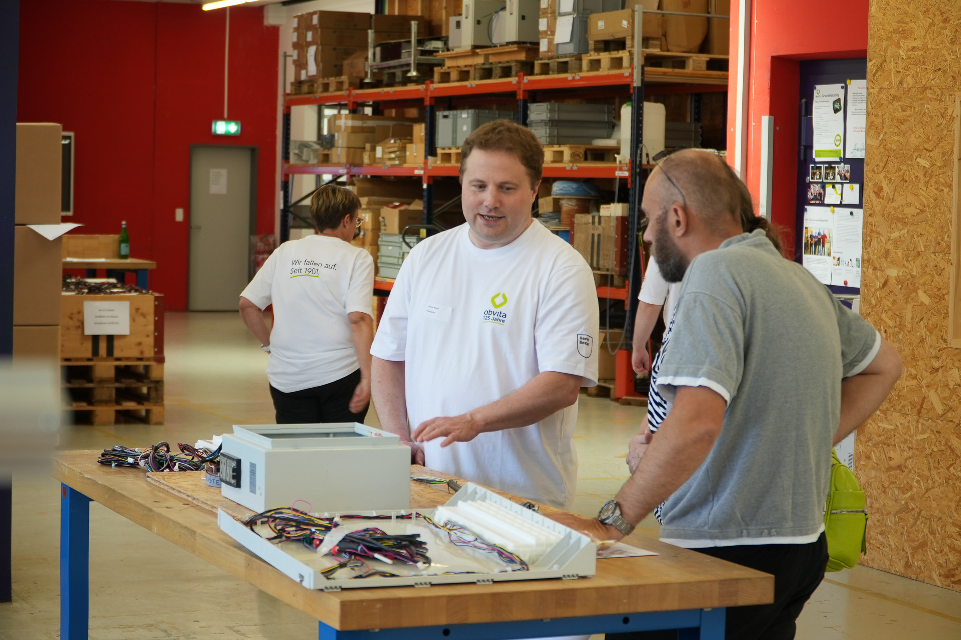 Zwei Männer stehen an einer Werkbank und unterhalten sich über elektronische Geräte in einer Lagerhalle, während im Hintergrund zwei weitere Personen arbeiten. Im Bild sind Regale mit Kartons und Bauteilen zu sehen.