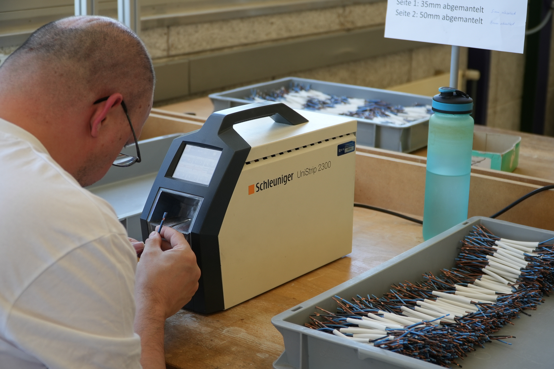 Eine Person bedient an einem Arbeitsplatz eine Schleuniger UniStrip 2300-Maschine und bereitet dabei in Schalen angeordnete Drähte vor; daneben steht eine Wasserflasche, und im Hintergrund sind Hinweisschilder zu sehen.