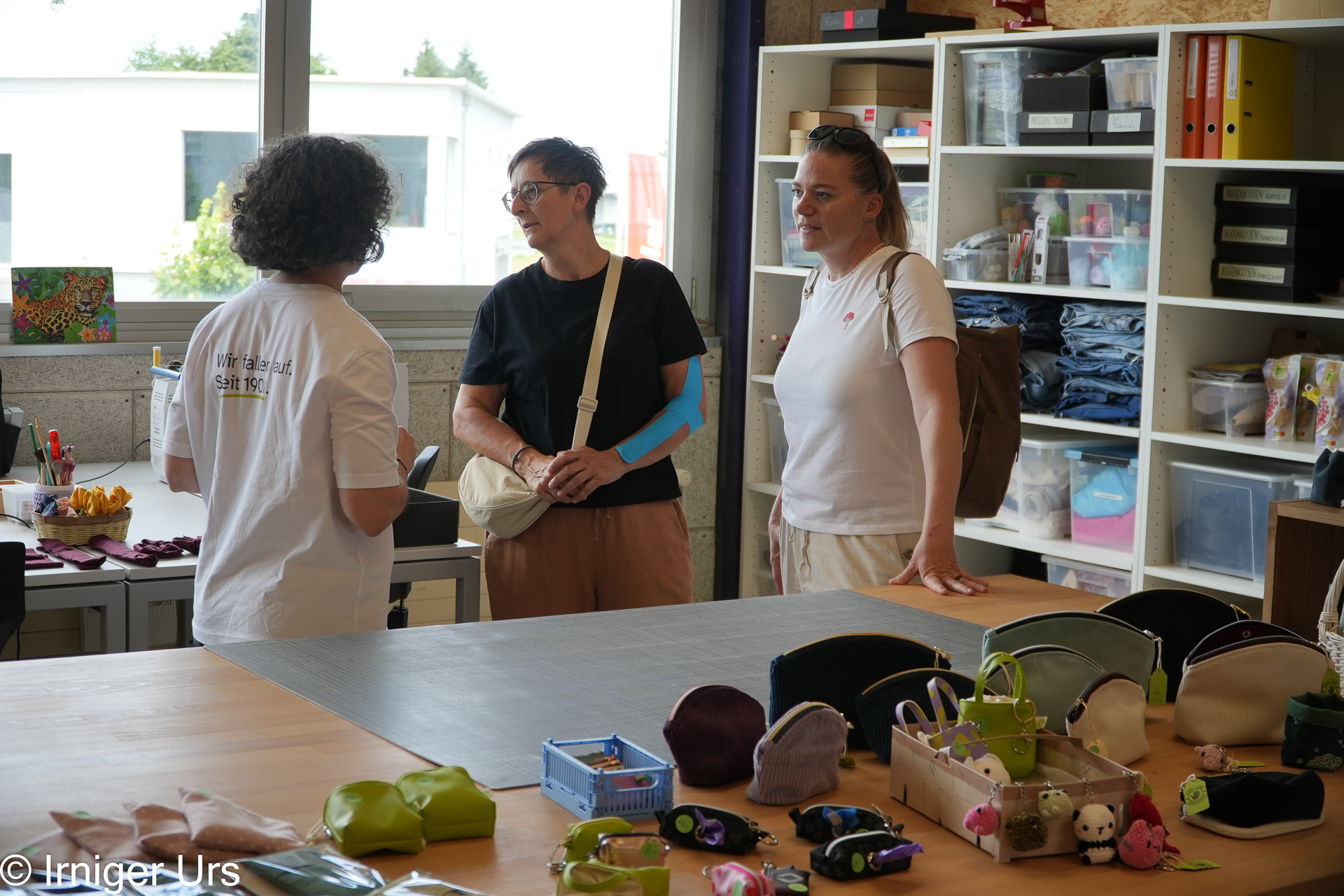Drei Frauen stehen in einer Werkstatt, die mit handgefertigten Beuteln, Spielzeug und Bastelmaterialien gefüllt ist, und unterhalten sich. Im Hintergrund sind Regale mit Stoffen und Materialien zu sehen, und durch ein Fenster fällt Sonnenlicht herein.