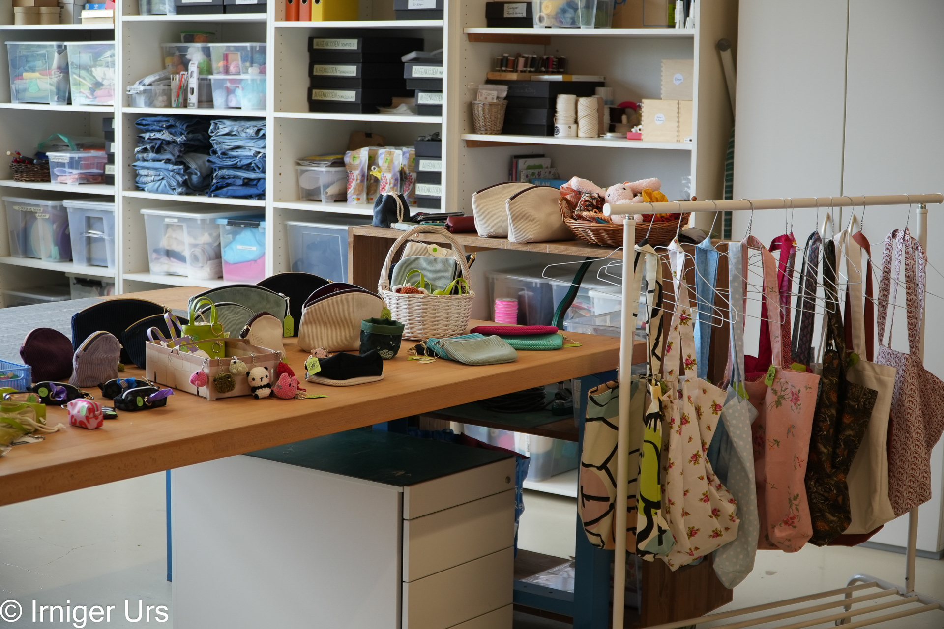 Eine Werkstatt mit Regalen voller Stoffe und Bastelmaterialien, ein Tisch, auf dem handgefertigte Beutel und kleine Bastelarbeiten ausgestellt sind, und daneben ein Kleiderständer, an dem bunte Tragetaschen und Beutel hängen.