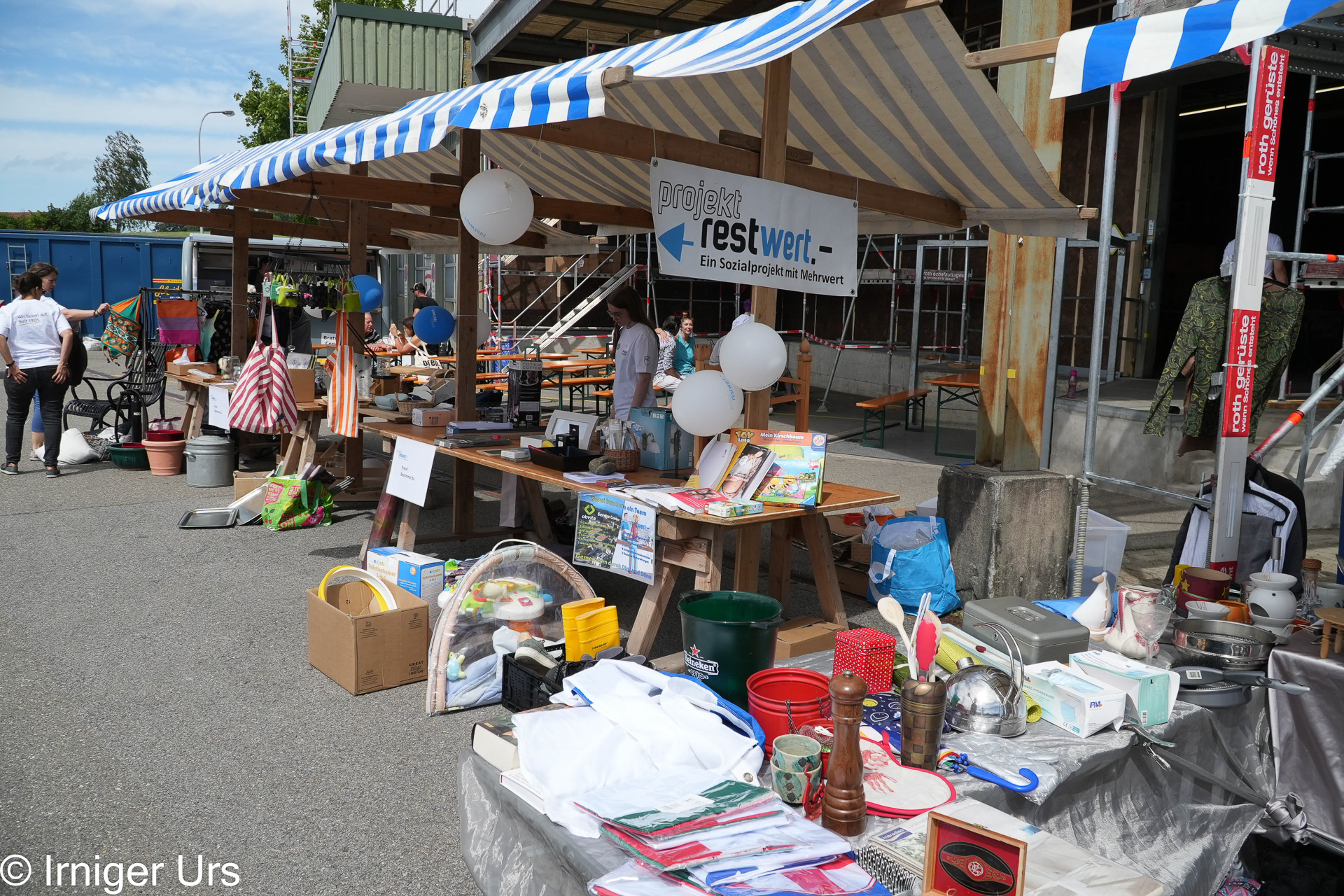 Ein Flohmarktstand mit verschiedenen Artikeln, darunter Bücher, Spielzeug, Küchenutensilien und Taschen. Blaue und weiße Luftballons sowie ein Banner mit der Aufschrift „projekt restwert“ schmücken den Holzstand. Im Hintergrund stöbern Menschen in den Waren.