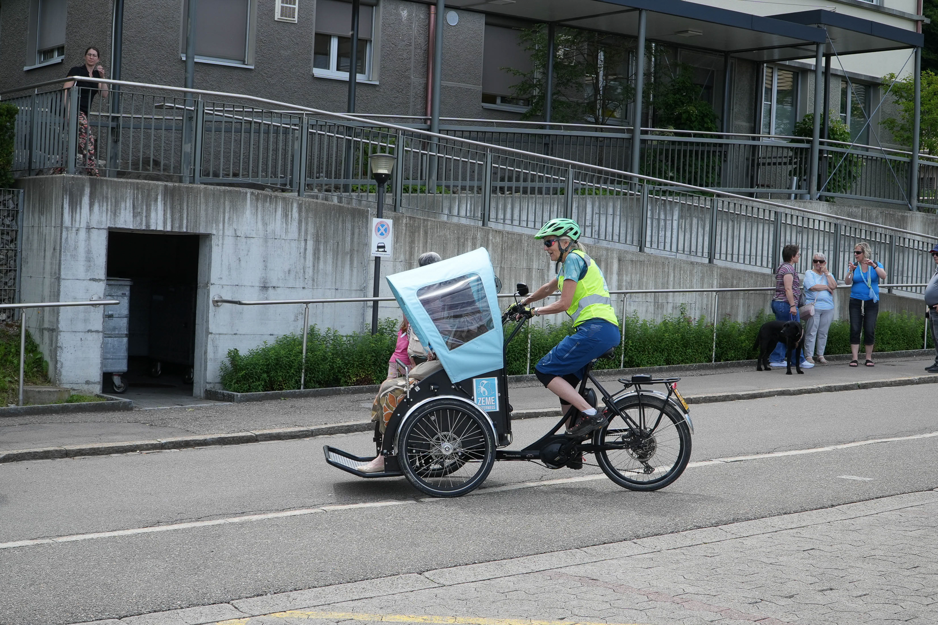 Eine Person, die einen Helm und eine Warnweste trägt, fährt mit einem Passagier auf dem Vordersitz ein Dreiradtaxi und fährt an einem Gebäude vorbei, während in der Nähe einige Menschen stehen und vorbeigehen.