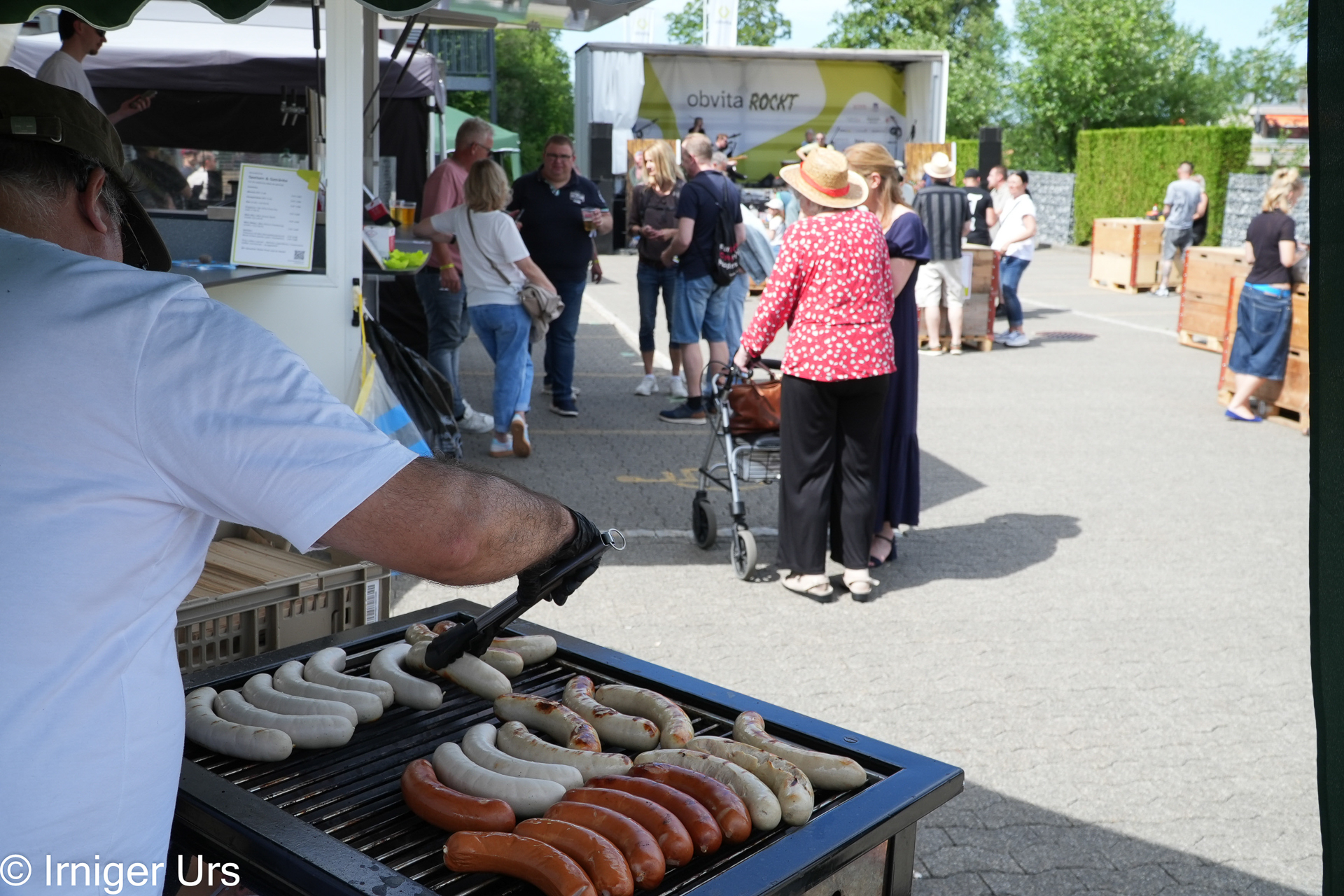 An einem sonnigen Tag grillt jemand auf einem Markt im Freien verschiedene Würstchen, während die Leute in der Schlange vor den Imbissständen stehen und sich unterhalten.