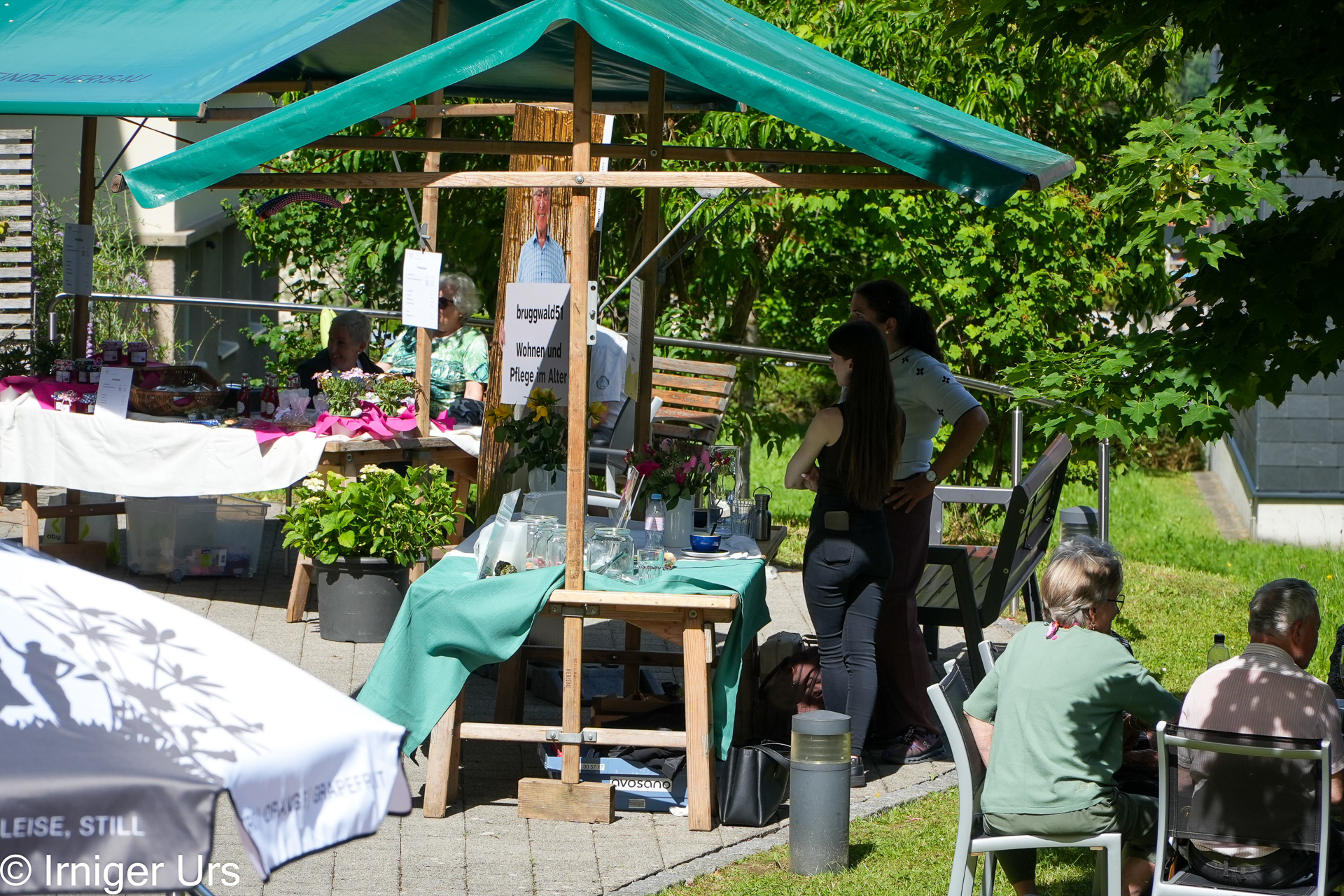 Die Menschen haben sich um die Stände eines Marktplatzes versammelt, die mit grünen Tischdecken gedeckt sind und im Schatten von Sonnenschirmen und Bäumen liegen. Einige sitzen an einem Tisch, während andere stehen und sich in der Nähe der Pflanzen- und Glasgefäßauslagen unterhalten. Sonnenlicht und Schatten vermischen sich in dieser Szene.