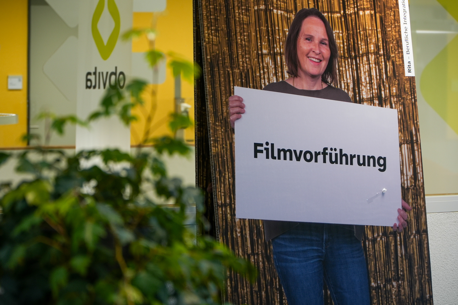 Eine Frau hält ein Schild mit der Aufschrift „Filmvorführung“ (auf Deutsch) in der Hand. Sie steht vor einer Kulisse mit Bambusmuster, im Vordergrund sind eine Pflanze und eine Glaswand mit der Aufschrift „obvita“ zu sehen.