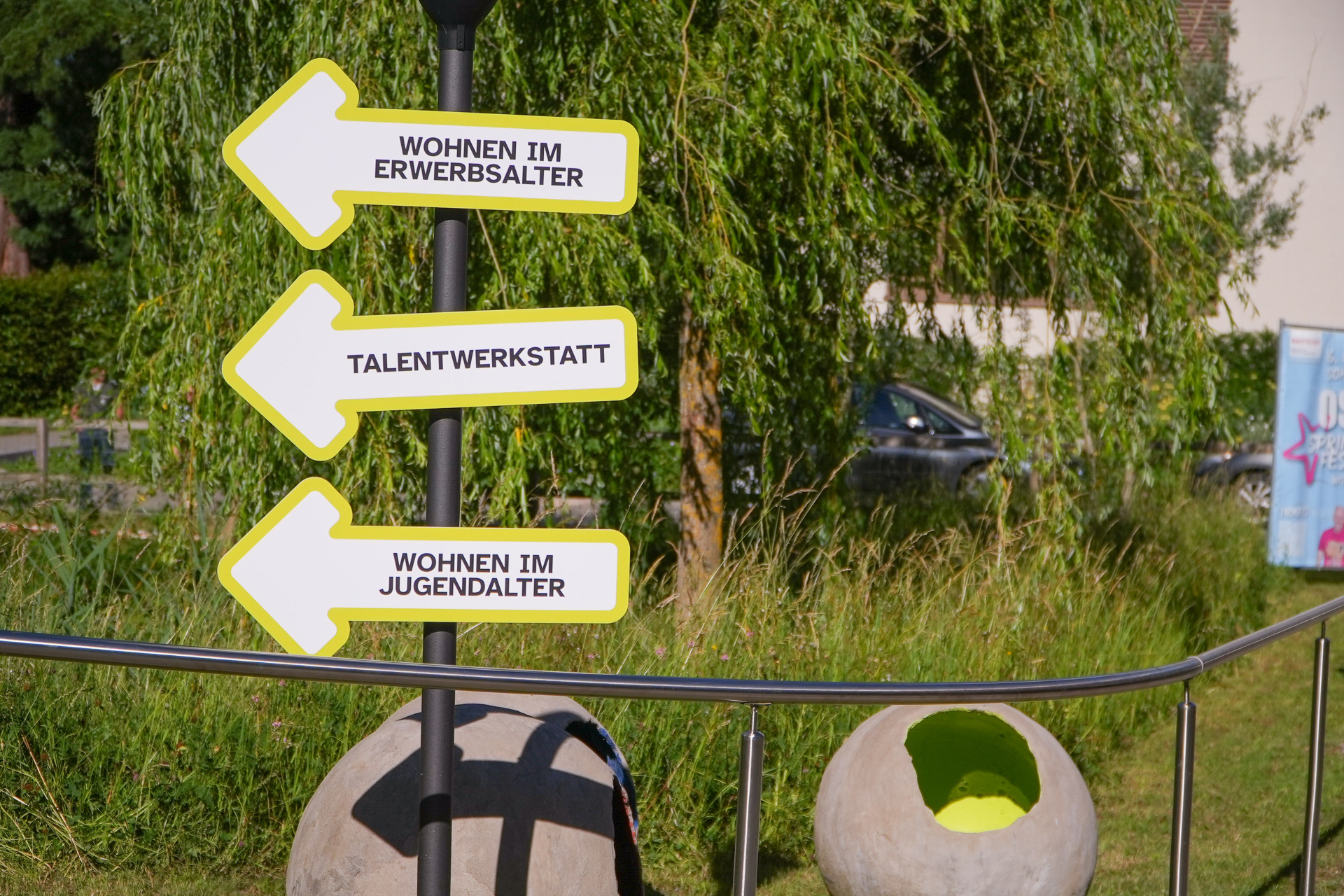 Auf einem Wegweiser mit drei gelben Pfeilen steht auf Deutsch: „Wohnen im Erwerbsalter“, „Talentwerkstatt“ und „Wohnen im Jugendalter“, im Hintergrund sind Grünflächen und große hohle Steinkugeln zu sehen.