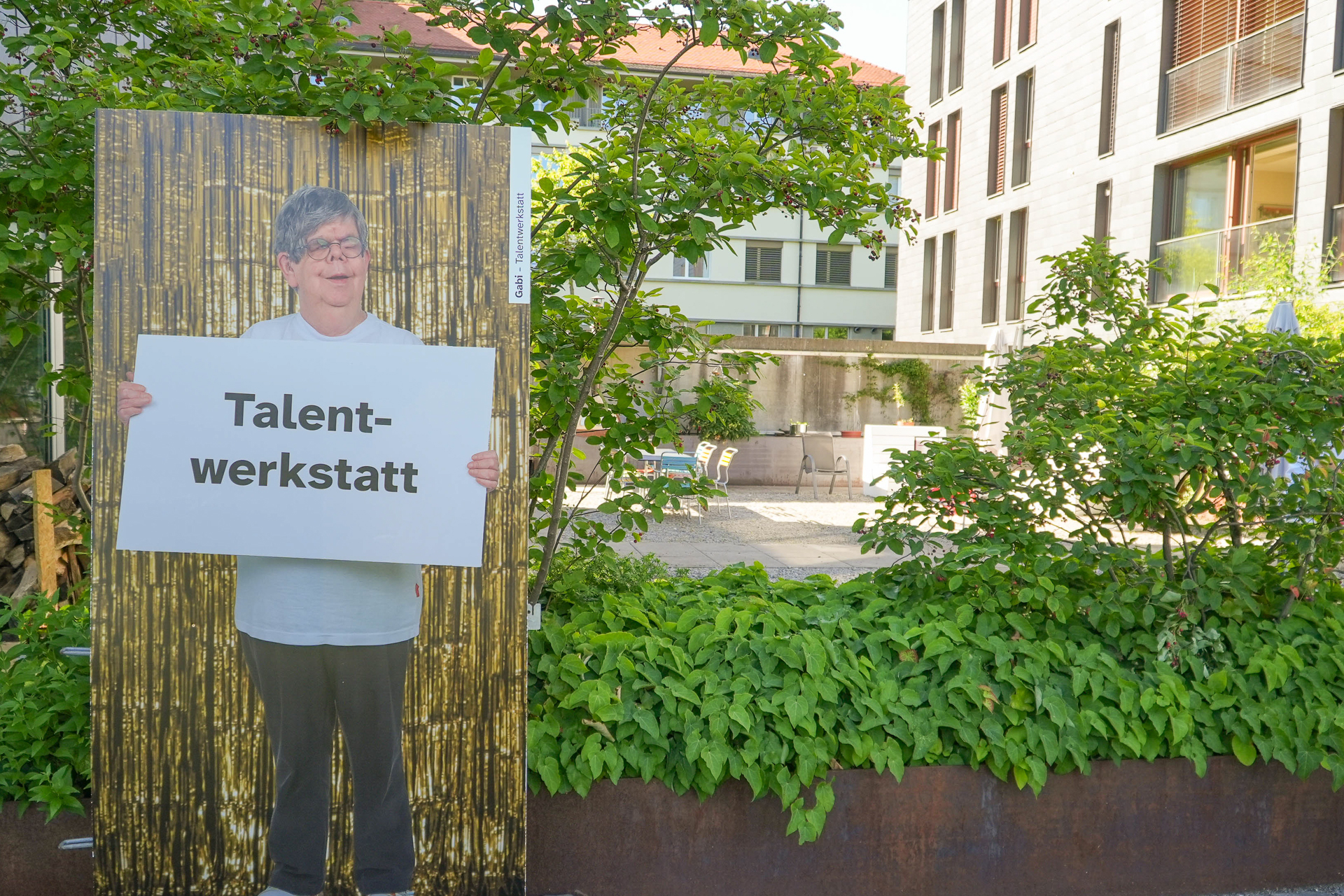 Im Freien, in einem grünen, von Bäumen gesäumten Innenhof neben modernen Gebäuden, steht eine lebensgroße Pappfigur einer älteren Person, die ein Schild mit der Aufschrift „Talent-werkstatt“ in der Hand hält.