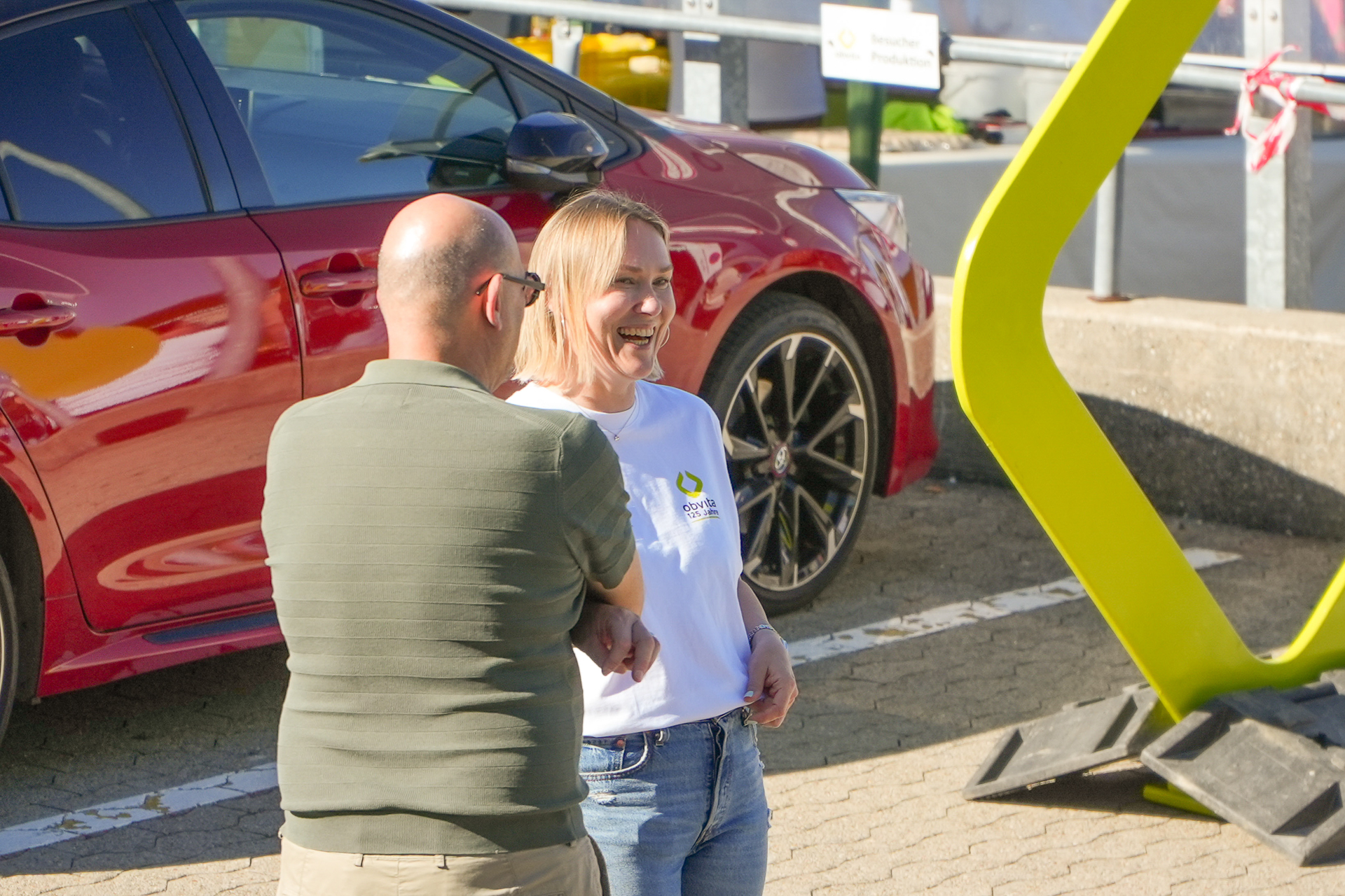 Ein Mann und eine Frau stehen auf einem sonnigen Parkplatz in der Nähe eines roten Autos und unterhalten sich; die Frau lächelt und trägt ein weißes Hemd, während der Mann in einem grünen Polohemd mit dem Rücken zur Kamera steht.