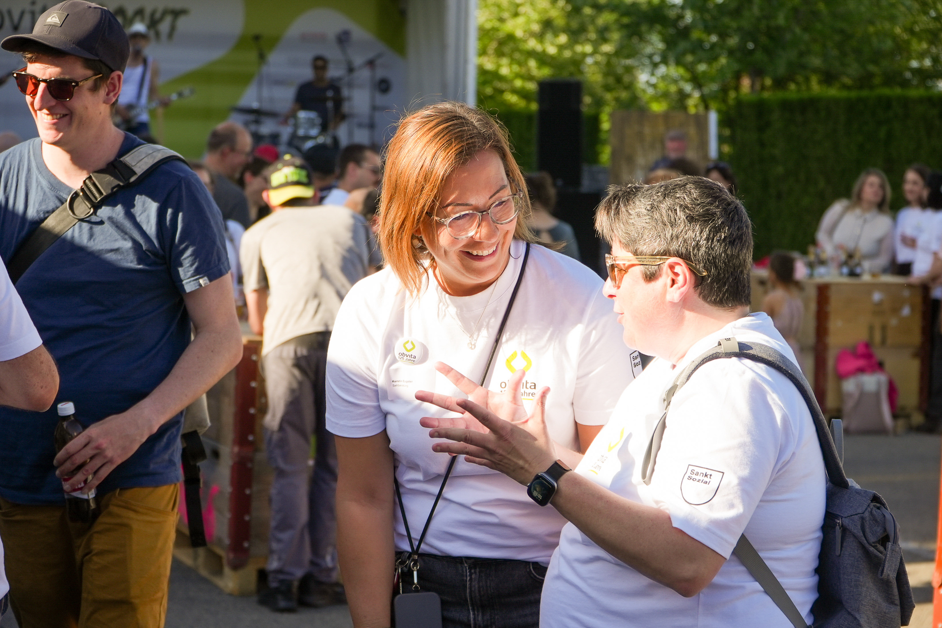 Zwei Menschen in weißen T-Shirts lächeln und unterhalten sich bei einer Veranstaltung im Freien, umgeben von anderen Teilnehmern. Der eine trägt eine Brille und hält ein Handy in der Hand, während der andere mit der Hand gestikuliert. Sonnenlicht taucht die Szene in helles Licht.