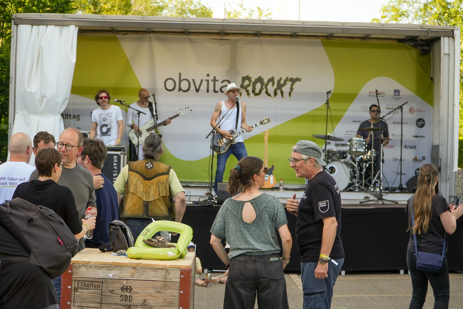 Auf einer mit „obvita ROCKT“ beschrifteten Außenbühne tritt eine Band auf, während sich die Besucher bei einer Tagesveranstaltung vor der Bühne unterhalten und zuschauen. Einige stehen, unterhalten sich oder sitzen in der Nähe der Technik.
