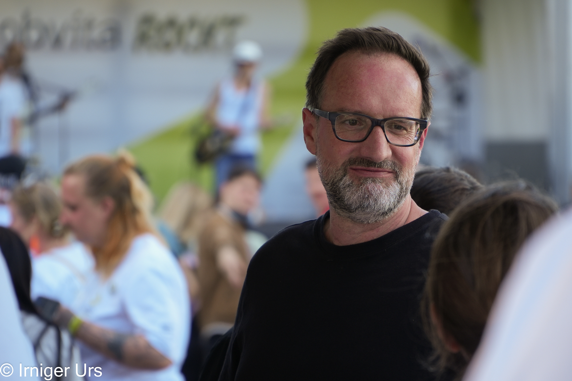 Ein Mann mit Brille und Bart lächelt, während er bei einer Veranstaltung im Freien inmitten einer Menschenmenge steht. Der Hintergrund ist leicht unscharf und zeigt Menschen sowie eine Bühne mit Musikern.