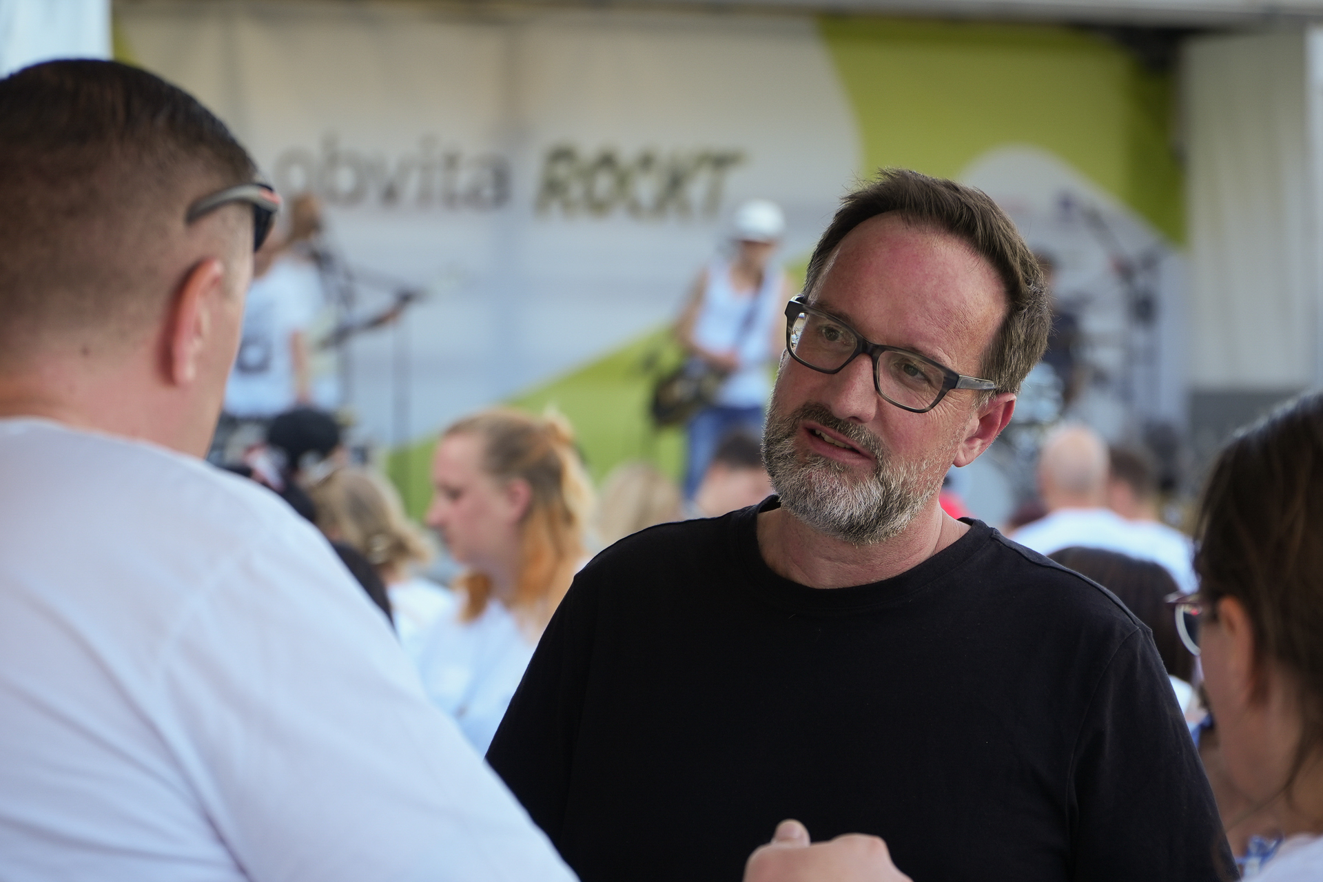 Ein Mann mit Brille und schwarzem Hemd unterhält sich bei einer Veranstaltung im Freien mit einer anderen Person, im Hintergrund sind eine verschwommene Menschenmenge und eine Bühne zu sehen. Auf dem Banner auf der Bühne steht „obvita ROCKT“.