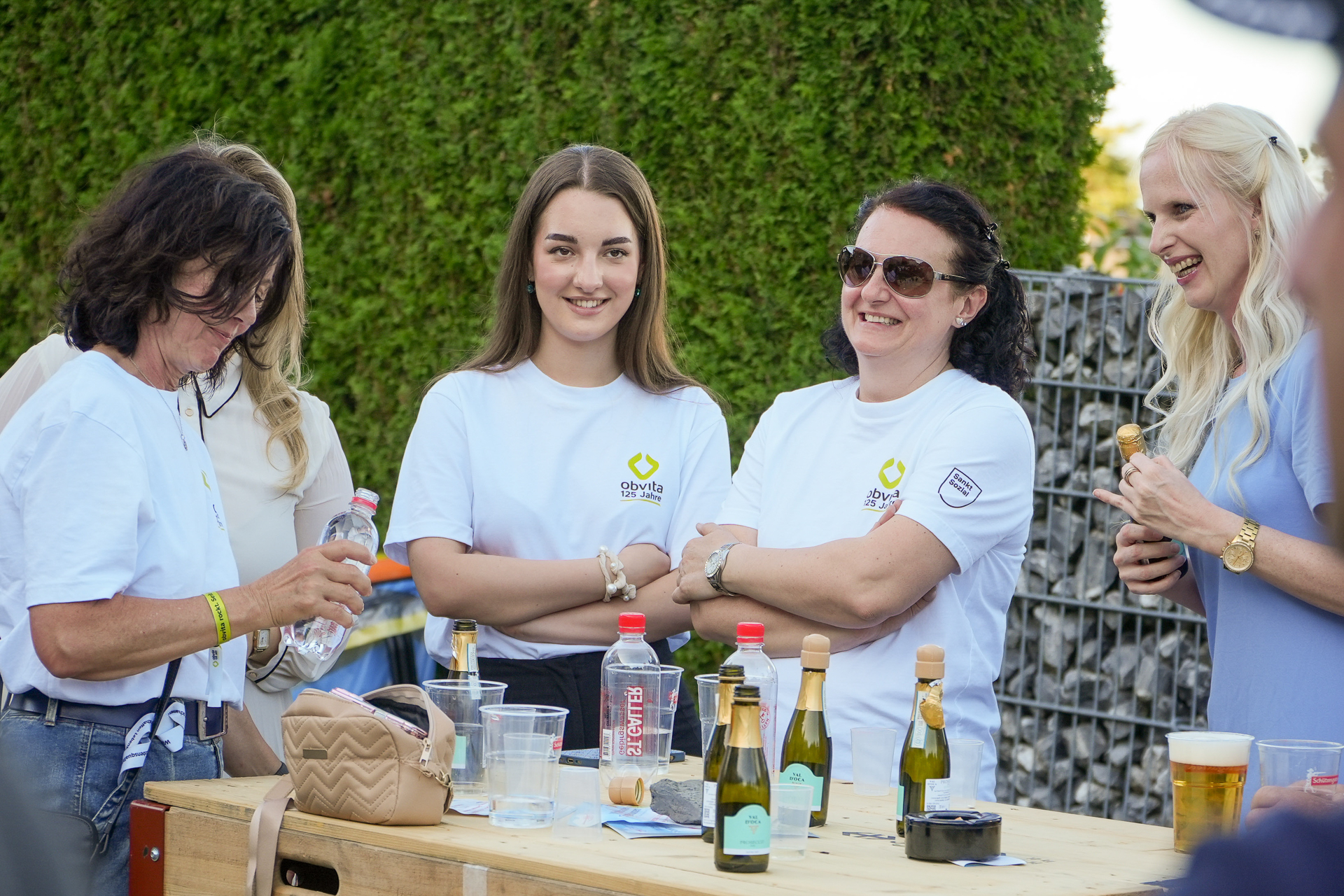 Vier Frauen stehen im Freien um einen Holztisch herum, lächeln und unterhalten sich. Auf dem Tisch stehen Getränke und Flaschen. Im Hintergrund sind grüne Büsche und ein Metallzaun zu sehen. Zwei Frauen tragen identische weiße Blusen.