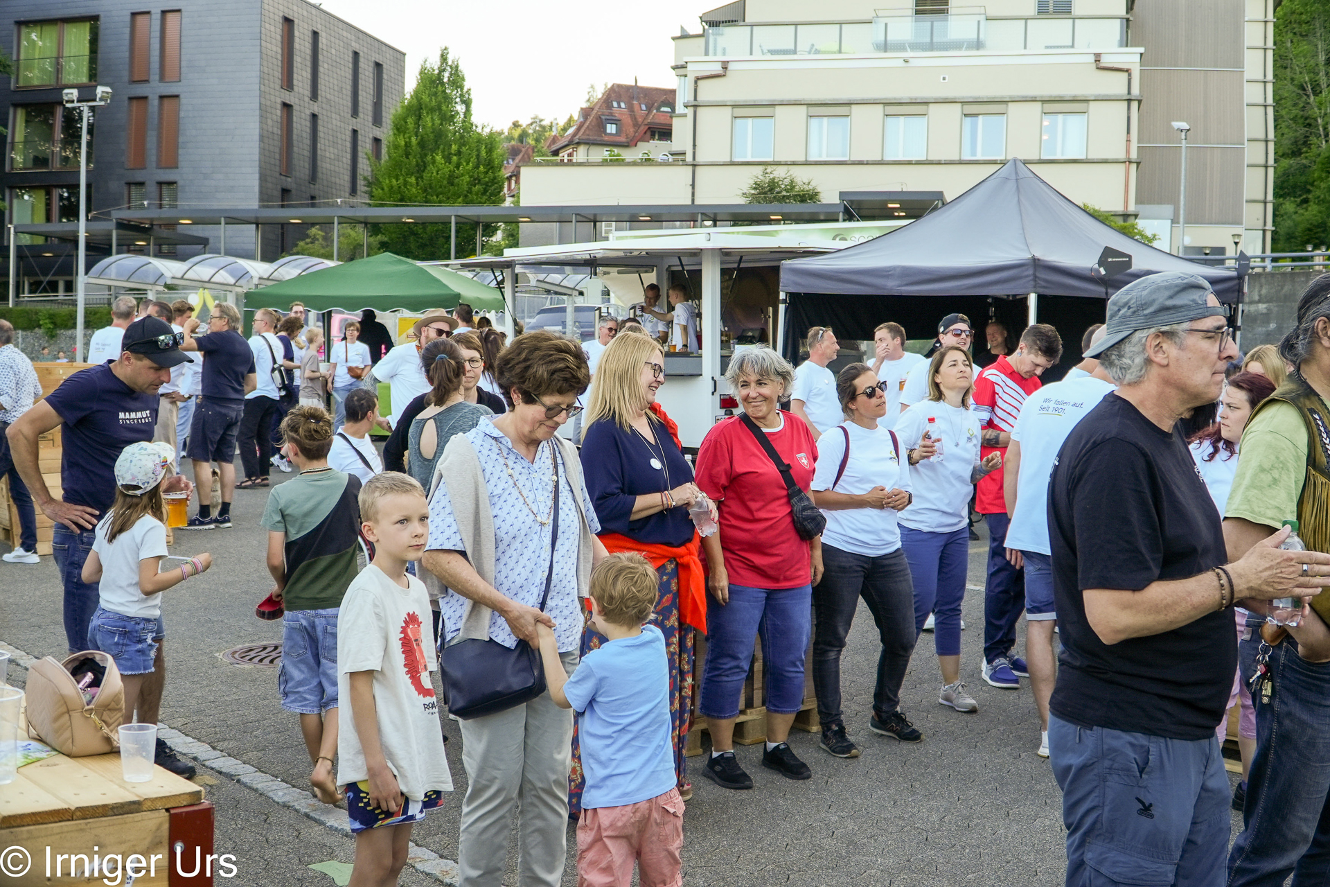An einem sonnigen Tag versammelt sich eine Menschenmenge, darunter Familien und Kinder, im Freien bei einer Straßenveranstaltung, bei der im Hintergrund Imbissstände und Zelte zu sehen sind. Einige unterhalten sich, während andere sich umschauen oder in der Schlange warten.