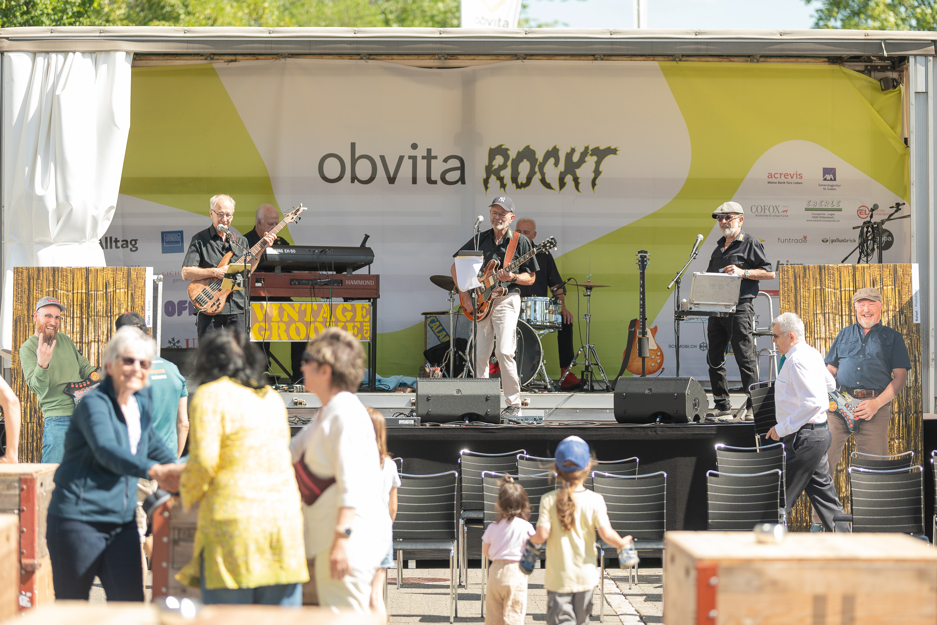 Auf einer mit „obvita ROCKT“ beschrifteten Außenbühne tritt eine Band auf, während im Vordergrund an einem sonnigen Tag Menschen zuschauen und sich unterhalten. Kinder und Erwachsene genießen die Veranstaltung.