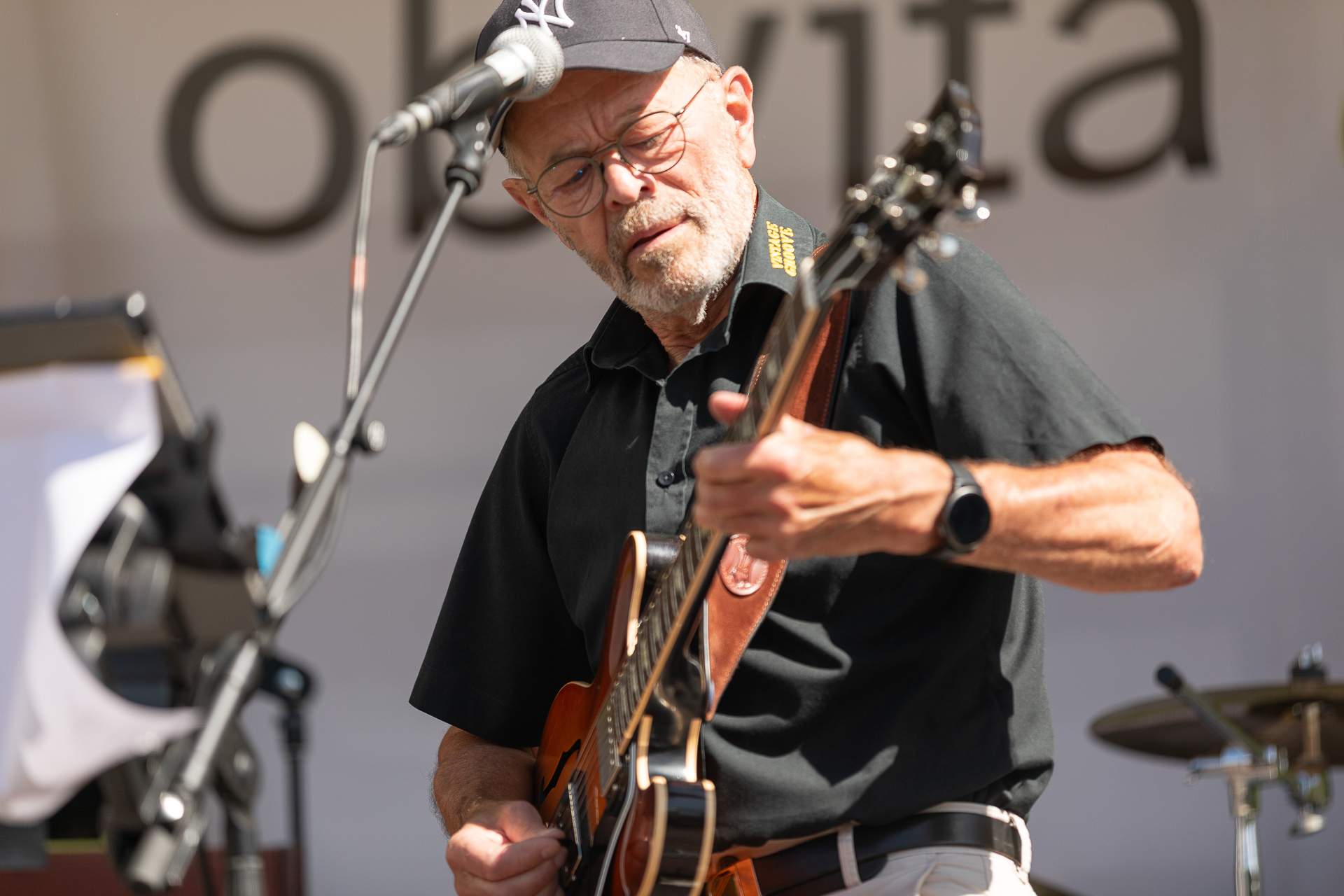 Ein älterer Mann mit Brille, schwarzem Hemd und Baseballmütze spielt auf einer Freiluftbühne E-Gitarre und singt ins Mikrofon, im Hintergrund sind verschwommene Instrumente und ein Schild zu sehen.