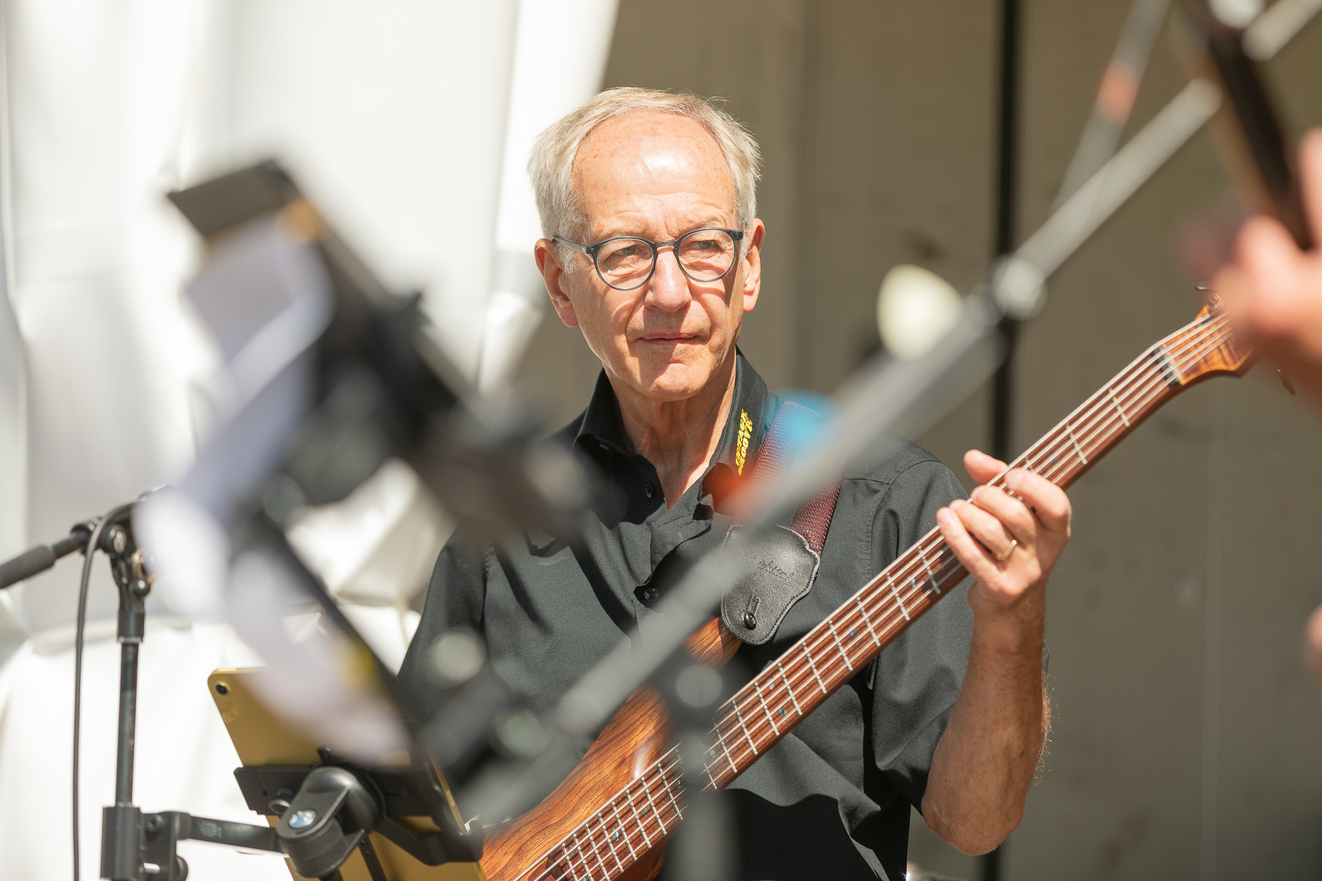 Ein älterer Mann mit Brille und schwarzem Hemd spielt im Freien E-Bass, während das Sonnenlicht sein Gesicht beleuchtet. Im Vordergrund sind Musikinstrumente und Mikrofonständer zu sehen.