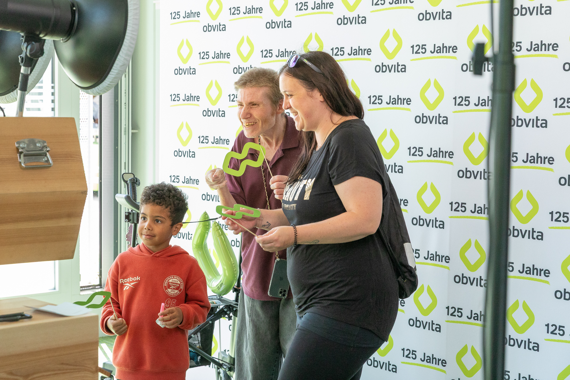 Drei Personen, darunter ein kleiner Junge und zwei Erwachsene, posieren bei einer Feier vor einem Hintergrund mit dem „obvita“-Logo und der Aufschrift „125 Jahre“ für ein Foto, wobei sie grüne Requisiten in den Händen halten.